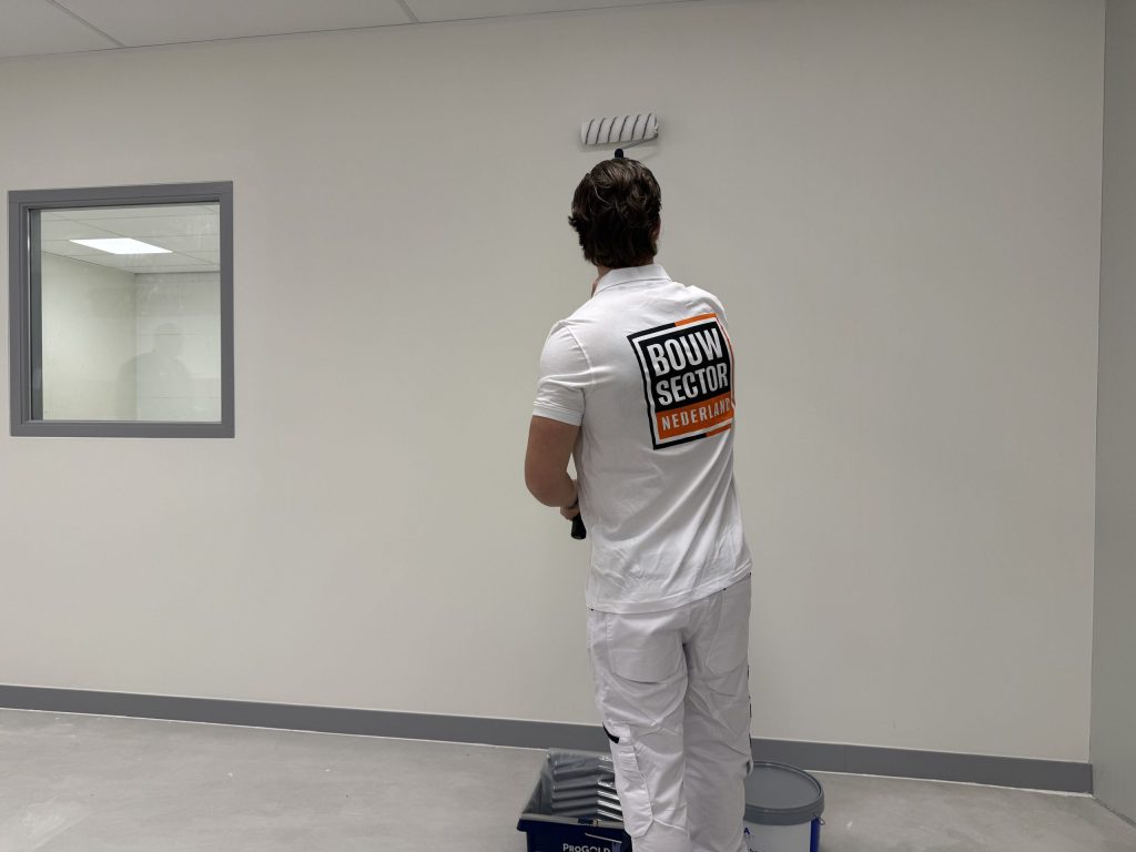 Schilder in een wit poloshirt verft een lichte muur naast een binnenraam met grijs kozijn