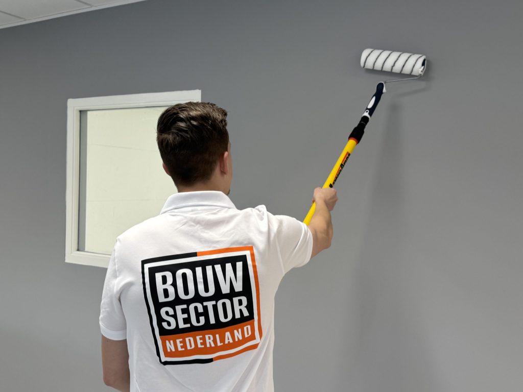 Schilder met het Bouwsector-logo op zijn rug zet grijze verf op de muur naast een raam met wit kozijn