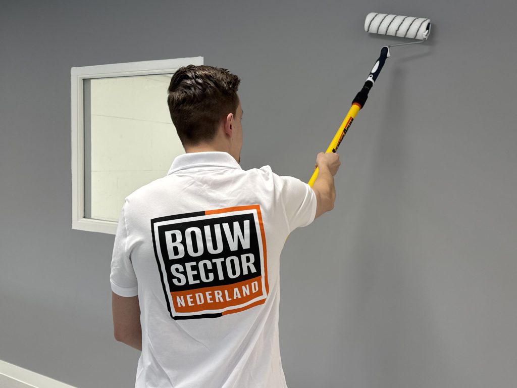 Schilder in witte werkpolo rolt een grijze muur vlak naast een wit binnenraam
