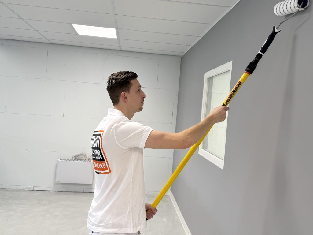 Schilder rolt een grijze muur bij een raam met een gele verlengsteel, met een radiator tegen de wand achter hem